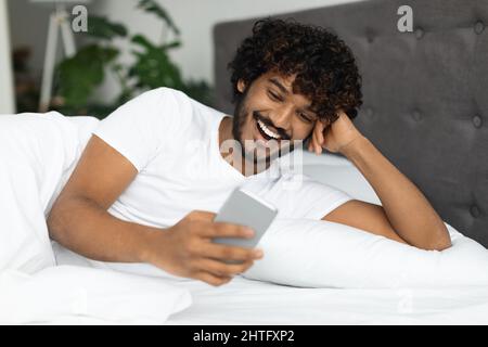 Glücklicher junger indischer Mann, der im Bett mit einer Freundin chattet Stockfoto