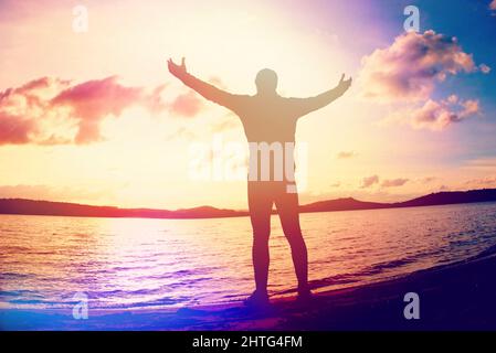 Mann, der am Ufer die Waffen hebt. Ein glücklicher Mann stand am Strand die Hände über den Kopf. Abstraktes Licht, farbenfrohe Lichtreflexe. Stockfoto