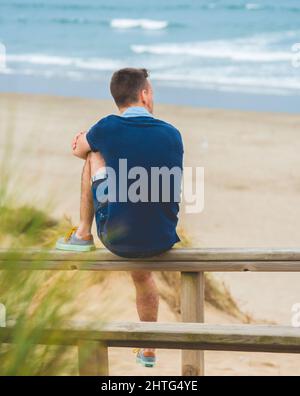 Vertikale Aufnahme eines Mannes, der auf dem Holzzaun sitzt und am Strand von Salin auf das Meer blickt Stockfoto