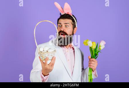 Ostern. Kaninchenmann in Hasenohren mit Blume. Bärtiger Rüde in Anzug hält Korb mit Eiern. Stockfoto