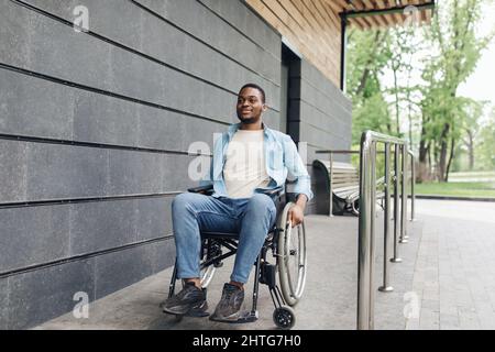 Glücklicher, tausendjähriger schwarzer Mann im Rollstuhl, der das Gebäude auf der Rampe verlässt und Einrichtungen für Menschen mit besonderen Bedürfnissen nutzt Stockfoto