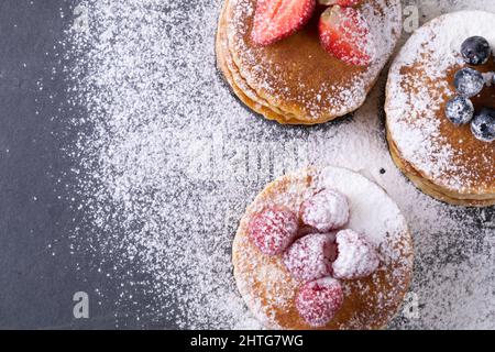 Direkt über Schuss Puderzucker über gestapelten Pfannkuchen mit verschiedenen Beerenfrüchten auf Futterschiefer Stockfoto