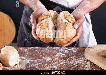 Nahaufnahme eines afroamerikanischen männlichen Bäckers, der gebackenes Brot über den Tisch brach Stockfoto