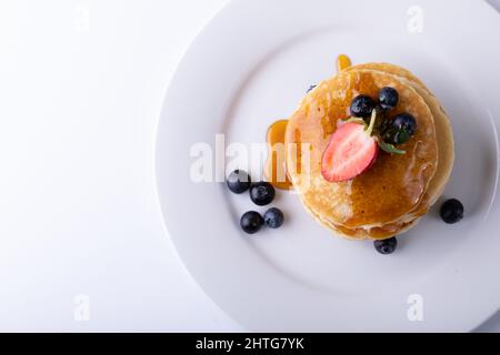Direkt über Schuss gestapelte Pfannkuchen mit Sirup und Beerenfrüchten in Teller auf weißem Hintergrund Stockfoto