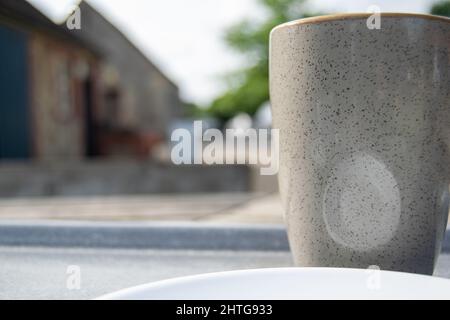 Nahaufnahme für eine grau gepunktete Kaffeetasse im Restaurant im Freien, Nahaufnahme mit einer Tasse auf der rechten Seite und verschwommenen Gebäuden dahinter. Stockfoto