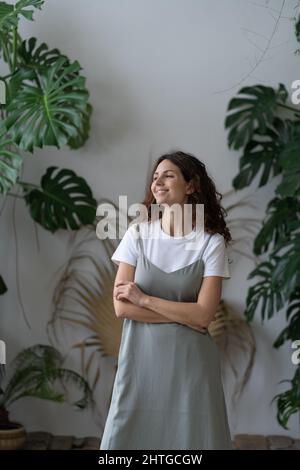 Junge glückliche weibliche Gärtnerin, die im Hausgarten gegen üppige Monstera Deliciosa Zimmerpflanzen steht Stockfoto