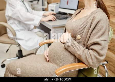 Ausgeschnittene Aufnahme einer Schwangeren jungen Frau, die obgyn in einer medizinischen Klinik mit Schwerpunkt auf Bauch, Kopierraum berät Stockfoto