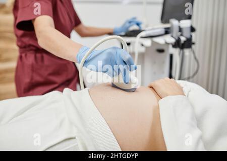 Nahaufnahme einer Ärztin, die den Ultraschallaufnehmer an den Bauch der Schwangeren hält, während sie die werdende junge Frau in der Klinik untersucht, Platz kopieren Stockfoto