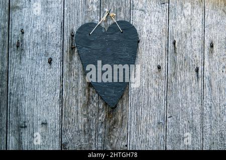 Schlichte schwarze Schieferherzenplatte auf einer Holzoberfläche mit verrosteten Nägeln, flache, herzförmige Skulptur aus Stein auf rustikalem Holz. Leerer Platz für den Kopiervorgang. Stockfoto