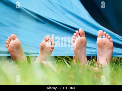 Barfuß auf einem Campingausflug. Blick aus der unteren Perspektive auf zwei Kinder, die barfuß unter einem Zelt liegen. Stockfoto