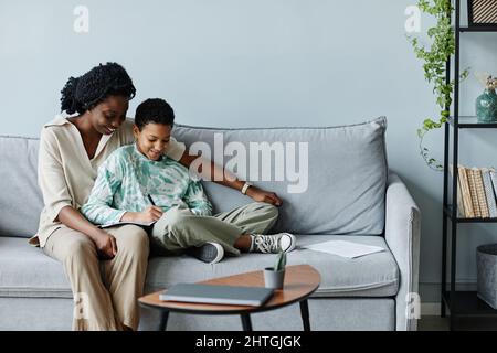 Minimales Porträt einer glücklichen schwarzen Mutter mit einem Teenager-Mädchen, das zusammen Hausaufgaben macht, während es zu Hause auf dem Sofa sitzt und Platz kopiert Stockfoto