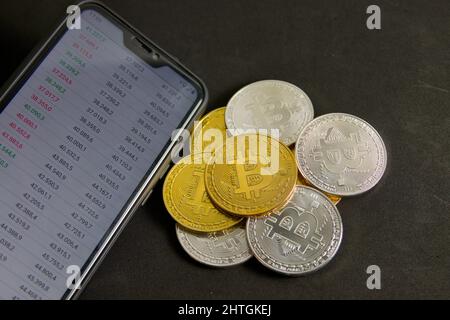 Nahaufnahme eines Stapels goldener und silberner Bitcoins neben einem Handy mit Wertetabellen auf schwarzem Hintergrund Stockfoto