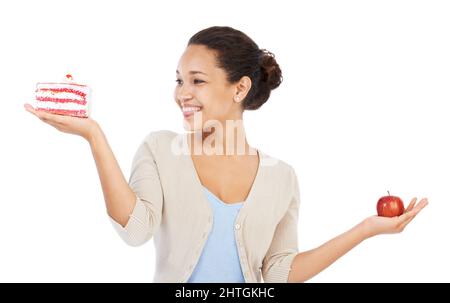 Ich habe definitiv diesen Kuchen. Junge Frau lächelt, während sie versucht, sich zwischen einem Stück Kuchen und einem Apfel zu entscheiden - isoliert auf Weiß. Stockfoto