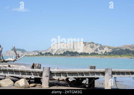 Driftwood-Baumstamm und Steg am schlammigen Rand des Uawa River, der durch die Gemeinde Tolaga Bay verläuft. Stockfoto