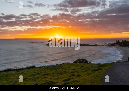 Goldenes Strahlen der Sonne, wenn es über dem fernen Horizont aufgeht und die Küste und den Off-Shore-Insel Mount Maunganui erhellt. Stockfoto