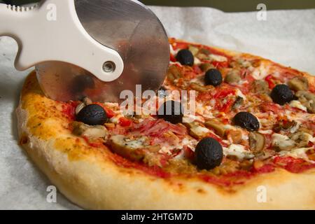 Eine hausgemachte Pizza mit Tomatensauce, Pilzen und schwarzen Oliven, die mit einem Cutter auf dem Ofenblech geschnitten werden Stockfoto