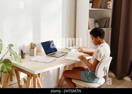 Minimales Porträt eines schwarzen Teenagers, das online in einem gemütlichen, von Sonnenlicht beleuchteten Wohninnenraum studiert Stockfoto