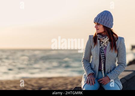 Frau in einem Wollmantel und Hut lächelt beim Sonnenuntergang, während sie den Horizont des Meeres betrachtet. Stockfoto