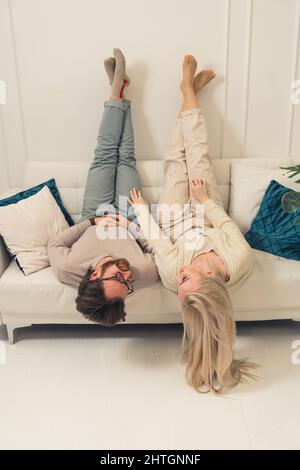 Junge kaukasische bärtige Unternehmer und seine schöne Freundin auf Sofa Beine nach oben vertikale drinnen Foto. Hochwertige Fotos Stockfoto