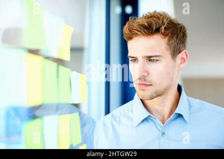 Nachdenklicher Typ, der sich Haftnotizen ansieht. Nachdenklicher Geschäftsmann, der sich Notizen auf Glas ansieht. Stockfoto