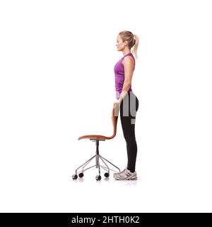 Stark stehen. Eine schöne junge Frau, die im Fitnessstudio Kleidung trägt und steht, während sie einen Stuhl vor weißem Hintergrund zur Unterstützung benutzt. Stockfoto