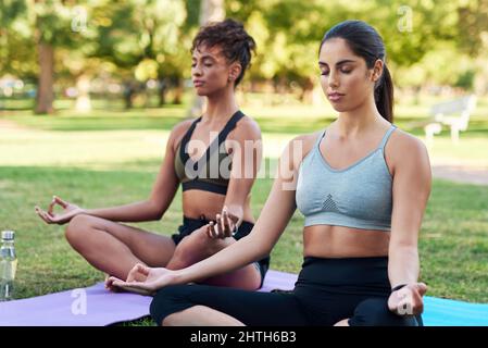 Sich auf Achtsamkeit und den Atem konzentrieren. Eine kurze Aufnahme von zwei attraktiven jungen Frauen, die nebeneinander sitzen und im Park meditieren. Stockfoto