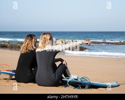 2 Surferfrauen sitzen am Strand und beobachten die Wellen Stockfoto