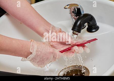 Frauenhände in Haarfärbehandschuhen waschen einen Kamm unter einem Wasserstrahl in einem weißen Waschbecken Stockfoto