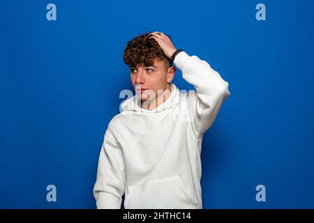 Junger, hübscher, großer, schlanker weißer Mann mit braunem Haar, das seinen Kopf mit Zweifelsgeste auf blauem Hintergrund kratzt. Stockfoto