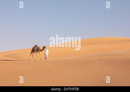 Mann in einer emiratischen Kandura, der mit einem Kamel auf den Wüstendünen im leeren Viertel spazierengeht. Abu Dhabi, VAE. Stockfoto