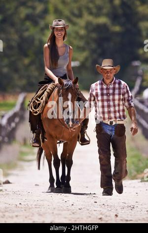Reiten 101. Ein attraktives junges Cowgirl, das auf einem Pferd auf einer Ranch reitet, während ein Cowboy an der Seite läuft. Stockfoto