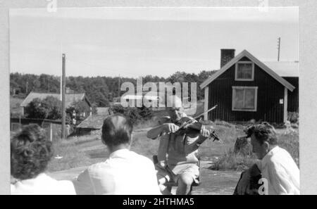 In den 1960er Jahren wurde ein Schwarz-Weiß-Foto des Mannes aufgenommen, der im Freien eine Geige spielt und von zwei Männern und einer Frau aus Schweden beobachtet wurde. Konzept der Freizeit, Entspannung, gestern Stockfoto