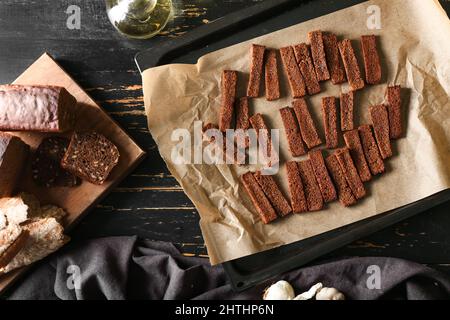 Backblech mit leckeren Roggencroutons und Brot auf schwarzem Holzhintergrund Stockfoto