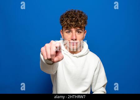 Junger, hübscher, dunkelhaariger Mann mit lockigen Haaren und weißem Sweatshirt auf blauem Hintergrund, der unzufrieden und frustriert auf die Kamera zeigt, wütend und Stockfoto