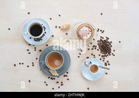 Vier Tassen mit verschiedenen Kaffee, Cappuccino, Espresso, Lungo schwarz und mit Crema, Cookies und gerösteten Bohnen auf einem hellen Holztisch, Kopierer Platz, hoch a Stockfoto