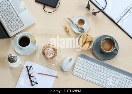 Von oben gesehen, mit Kaffeetassen und Keksen, Laptop, Tastatur, Ordner und Papieren auf hellem Holzhintergrund, niemand, ausgewählter Fokus Stockfoto