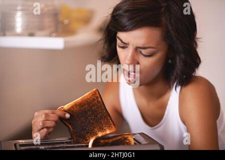 Oh nein Ein kurzer Schuss einer jungen Frau, die ein Stück verbranntem Toast aus dem Toaster nahm. Stockfoto