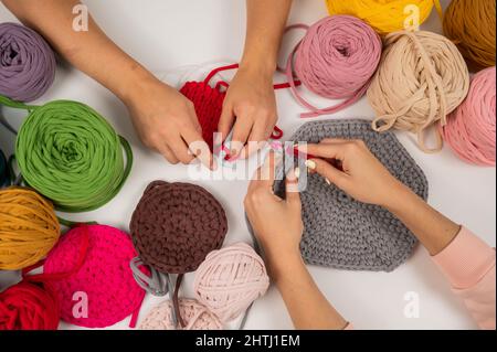 Draufsicht auf weibliche Hände mit Stricken auf einem weißen Tisch. Zwei Frauen stricken Körbe aus Baumwollgarn. Stockfoto