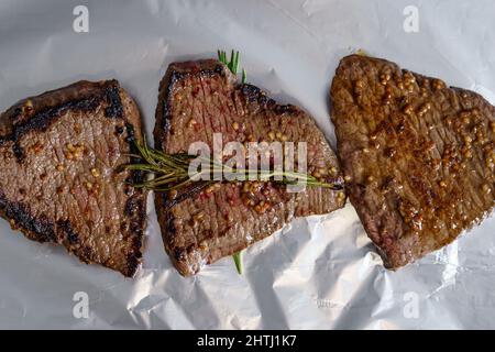 Drei geröstete Rindersteaks mit Rosmarin- und Senfkörnern auf Folie isoliert. Gegrilltes Fleisch. Draufsicht Stockfoto