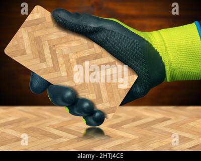 Nahaufnahme einer Hand mit schützenden Arbeitshandschuhen, die eine Holzparkettprobe aus Fischgrätholz auf einem ähnlichen Boden halten, mit Schatten und Reflexionen. Stockfoto