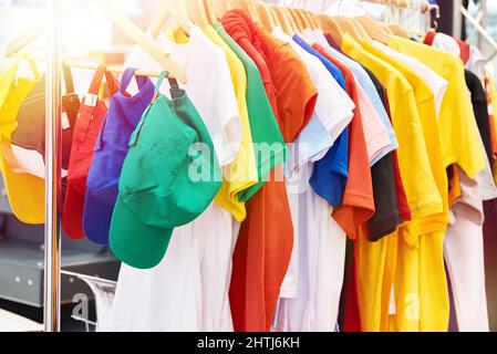 Farbige T-Shirts und Kappen auf dem Kleiderbügel im Laden Stockfoto