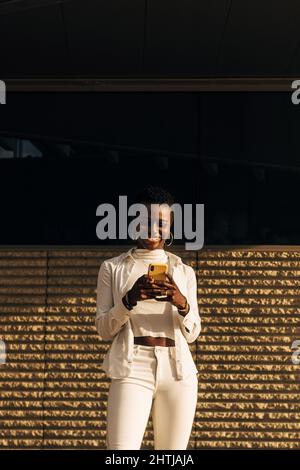 Stilvolle afroamerikanische Frau lächelt, während sie ihr Mobiltelefon im Freien benutzt. Stockfoto