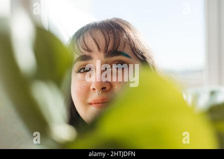 Positives junges Weibchen, das die Kamera anschaut und das Gesicht mit einem grünen Monstera-Blatt bedeckt, während es zu Hause im hellen Raum mit Sonnenlicht steht Stockfoto