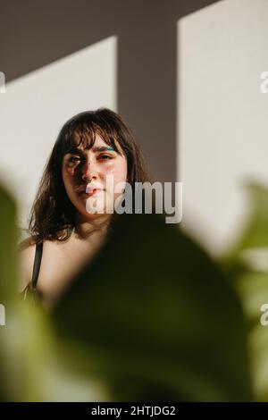 Positives junges Weibchen, das die Kamera anschaut und das Gesicht mit einem grünen Monstera-Blatt bedeckt, während es zu Hause im hellen Raum mit Sonnenlicht steht Stockfoto