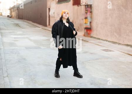 Junge Frau mit grün gefärbten Haaren, die eine Mütze und eine schwarze Jacke trägt und auf die Kamera schaut, während sie auf der Straße mit Gebäuden auf der sonnigen Straße steht Stockfoto