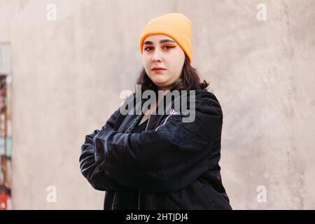 Junge Hündin mit grün gefärbtem Haar und einer Mütze in schwarzer Jacke, die in der Nähe einer orangefarbenen Wand auf einer sonnigen Straße auf die Kamera schaut Stockfoto