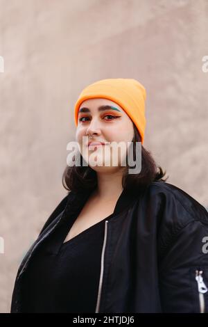 Junge lächelnde Frau mit grün gefärbten Haaren, die eine Mütze und eine schwarze Jacke trägt und die Kamera anschaut, während sie in der Nähe einer Wand auf einer sonnigen Straße steht Stockfoto