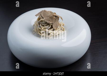 Appetitliche Pasta mit Sauce und leckeren schwarzen Trüffeln, serviert auf weißem runden Teller auf schwarzem Tisch in hellem, modernen Restaurant Stockfoto