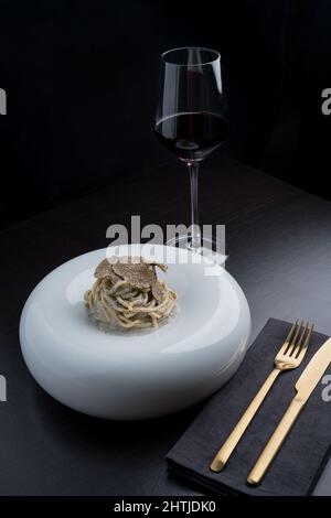 Appetitliche Pasta mit Sauce und leckeren schwarzen Trüffeln, serviert auf weißem runden Teller auf schwarzem Tisch neben goldenem Besteck in hellem, modernem Restaurant Stockfoto