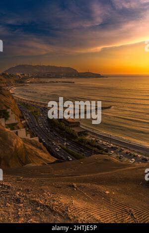 Sandiger Hang über der Straße mit Autos, die zur Sonnenuntergangszeit an der Küste entlang des plätschernden Meeres gegen den bewölkten Himmel in Lima fahren Stockfoto